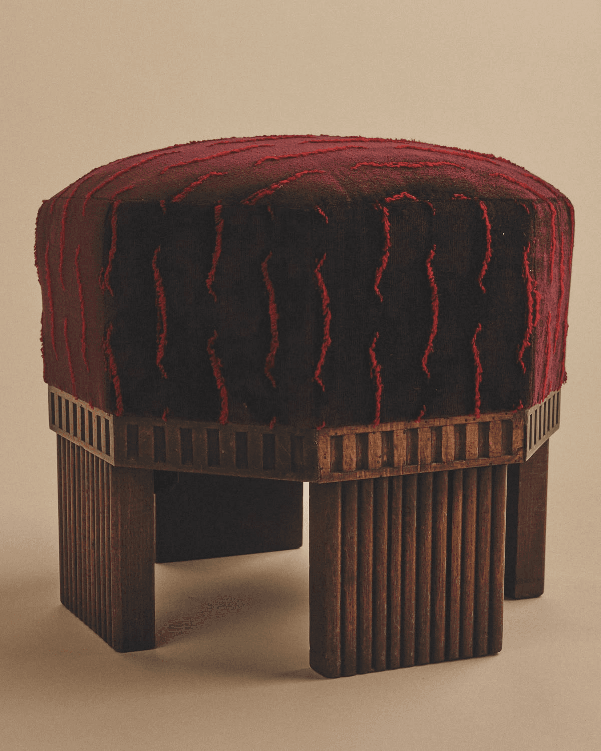 An Art Deco style upholstered stool featuring a hexagonal top with rich, deep red fabric that has a contrasting lighter red textured, wavy pattern. The stool is supported by a dark wooden base with a distinctive frieze of rectangular cutouts just below the upholstery, transitioning to fluted or reeded vertical panels that form the legs. — 3D model