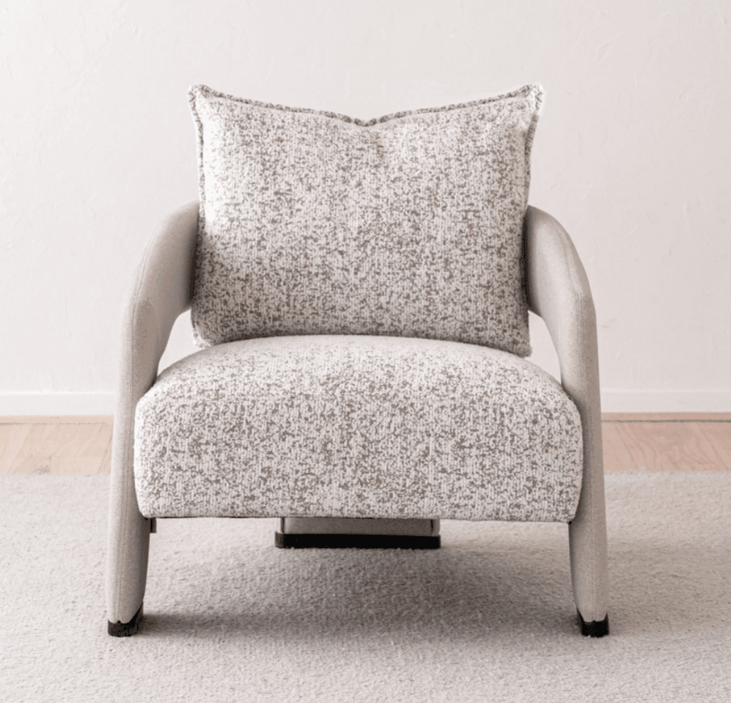A contemporary upholstered armchair featuring a rounded silhouette and a neutral-toned, speckled fabric. The chair includes integrated, curved armrests and an upholstered backrest, complemented by a separate, plush back cushion with piped edges. The seat appears integrated, and the chair stands on short, upholstered legs or a low, concealed base, suitable for modern interior settings. — 3D model