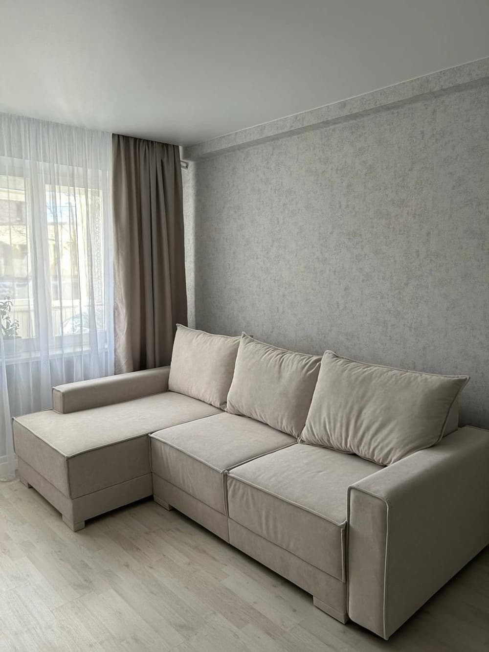 An L-shaped sectional sofa upholstered in a light beige fabric, featuring an integrated left-hand facing chaise lounge. The sofa has a clean, modern aesthetic with visible stitching details, low-profile block feet, and three separate back cushions. The armrests are broad and flat, matching the overall minimalist silhouette. — 3D model
