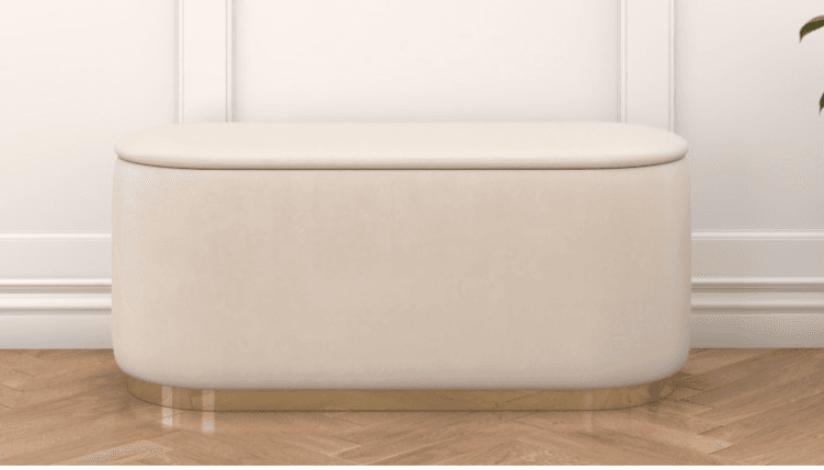 A rectangular ottoman with rounded corners, upholstered in a soft, cream-colored fabric. It features a padded top that appears to be a lift-off lid, suggesting internal storage. The base is accented with a polished gold-toned metal band that wraps around the bottom perimeter. — 3D model