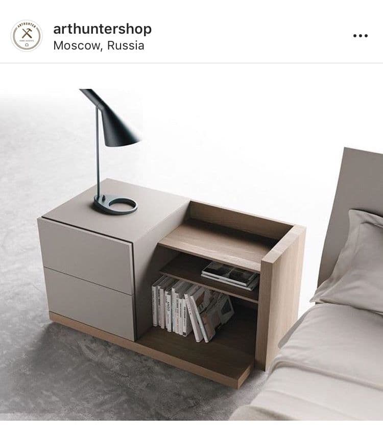 A modern, asymmetric nightstand featuring a two-drawer cabinet section in light gray and an integrated, lower open shelving unit in light wood. A small black conical table lamp rests on the top surface of the drawer unit, which is part of a wider base. — 3D model