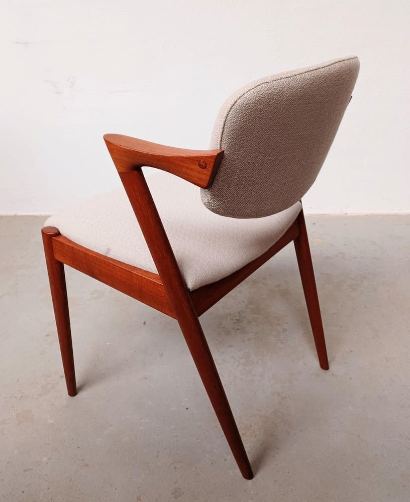 A mid-century modern armchair with a teak wood frame and upholstered seat and backrest. The frame features a distinctive angular design with tapered legs and elegantly curved armrests. The upholstery is a textured, light-colored fabric. — 3D model