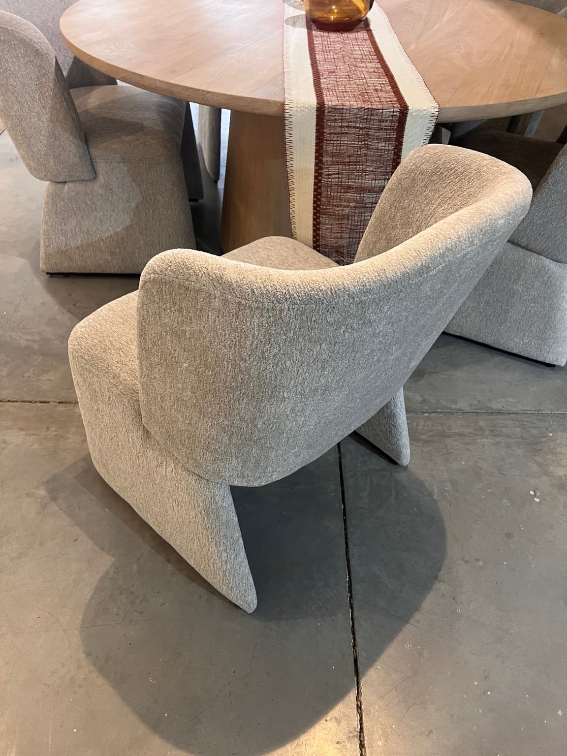 An upholstered dining chair featuring a contemporary sculptural form, integrated arms, and a continuous silhouette. It is covered in a light, textured, boucle-like fabric and is positioned at a dining table. — 3D model