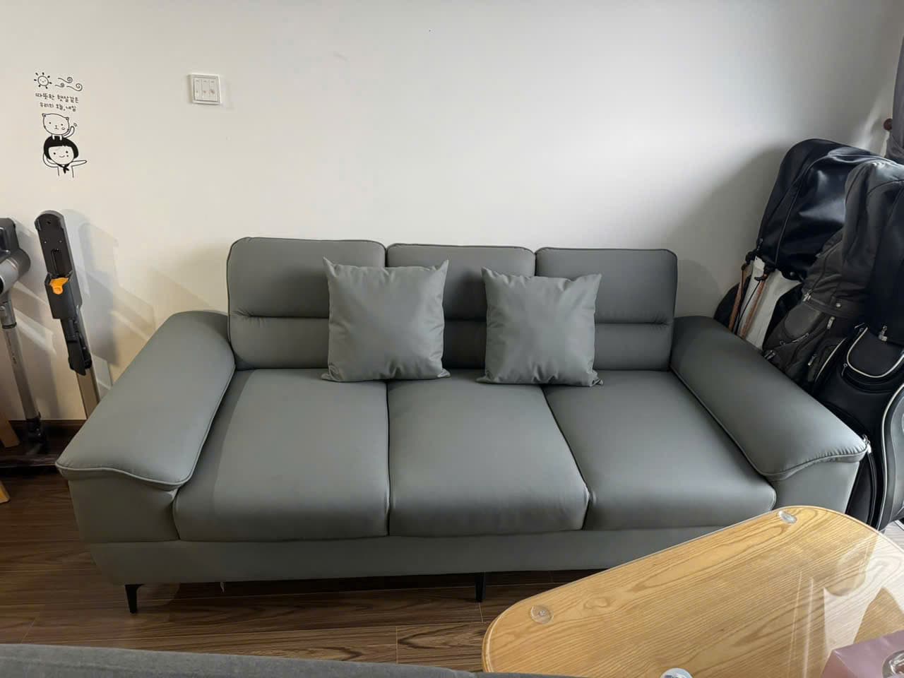 A modern three-seater sofa rendered in a neutral grey tone. It features clean lines, broad padded armrests with a slight curve, and three individual seat and back cushions. Two matching square throw pillows are also present. The sofa is supported by four slender, tapered black legs, characteristic of contemporary design. — 3D model