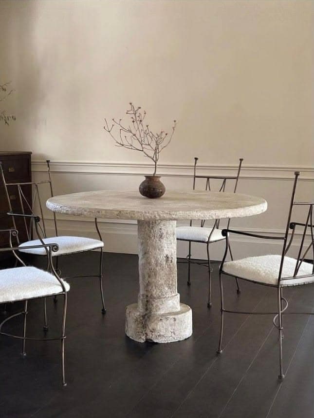 A round, pedestal table with a rough, textured surface. The pedestal has a columnar design with a decorative base and capital, reminiscent of classical architecture. A small, dark ceramic vase with a dried plant is placed on the tabletop. — 3D model