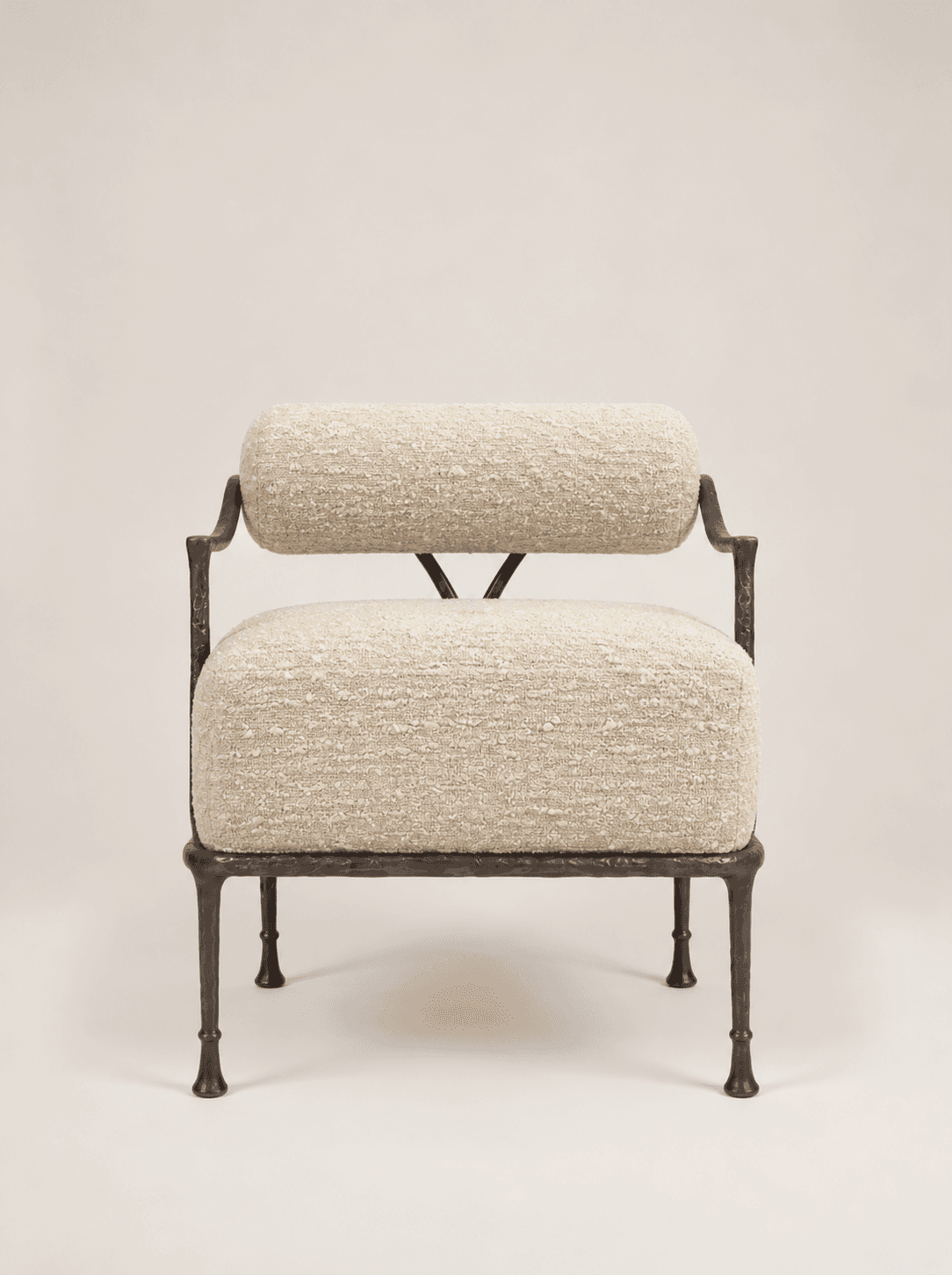An accent chair featuring a dark, hammered or forged metal frame and upholstered in a light-colored, textured boucle-like fabric. The design includes a separate cylindrical bolster for the backrest and a rectangular cushion for the seat. The metal frame forms minimal armrests and sturdy, tapered legs with small feet. The overall aesthetic is modern and artisanal. — 3D model