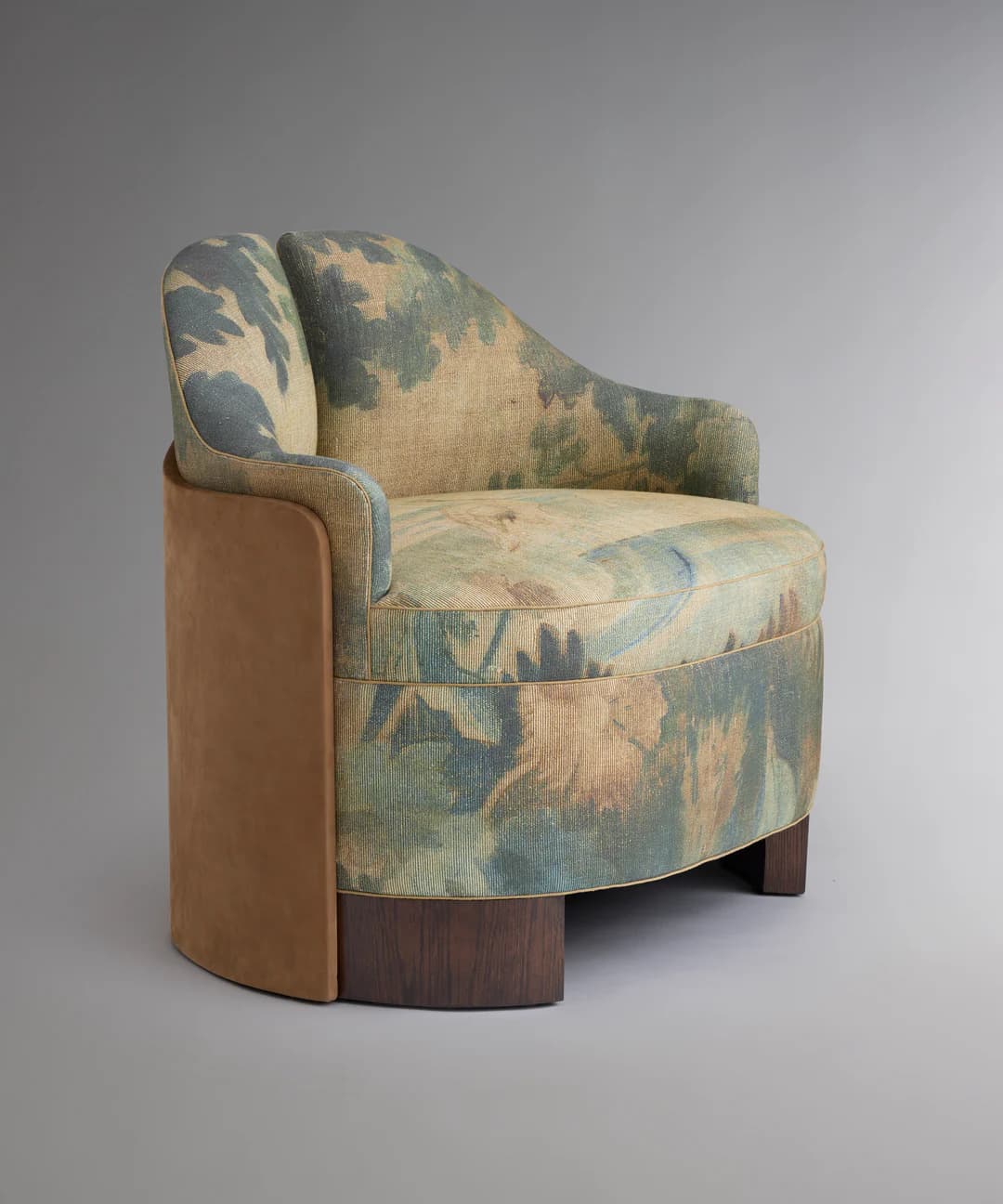 A single armchair with a rounded, upholstered body. The main upholstery features a large-scale floral or botanical pattern in muted greens, blues, and beige tones. The sides of the chair are covered in a contrasting smooth, tan material, possibly suede or a similar textile. The base is a dark wood, possibly walnut or mahogany, with a curved, pedestal-like design. The back of the chair has a subtle wingback element. — 3D model