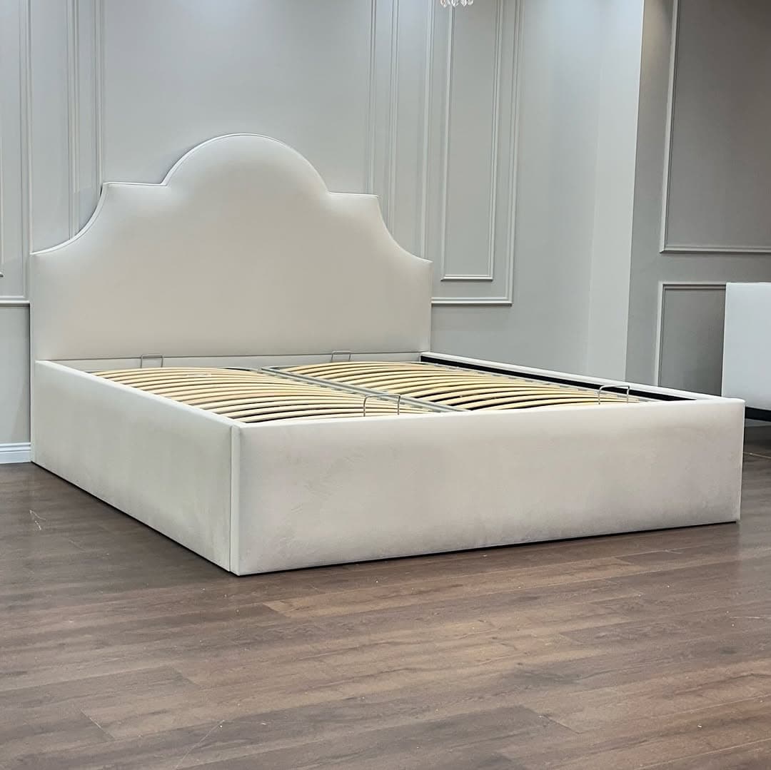 A contemporary upholstered platform bed frame featuring a distinctive scalloped headboard and a robust slatted base. The entire frame, including the headboard and side rails, is covered in a light-colored fabric, suggesting a soft and inviting aesthetic. The bed base incorporates a lift-up mechanism, indicated by the metal handles and exposed hardware, revealing under-bed storage space. The wooden slats are arranged to provide optimal support for a mattress without the need for a box spring. — 3D model