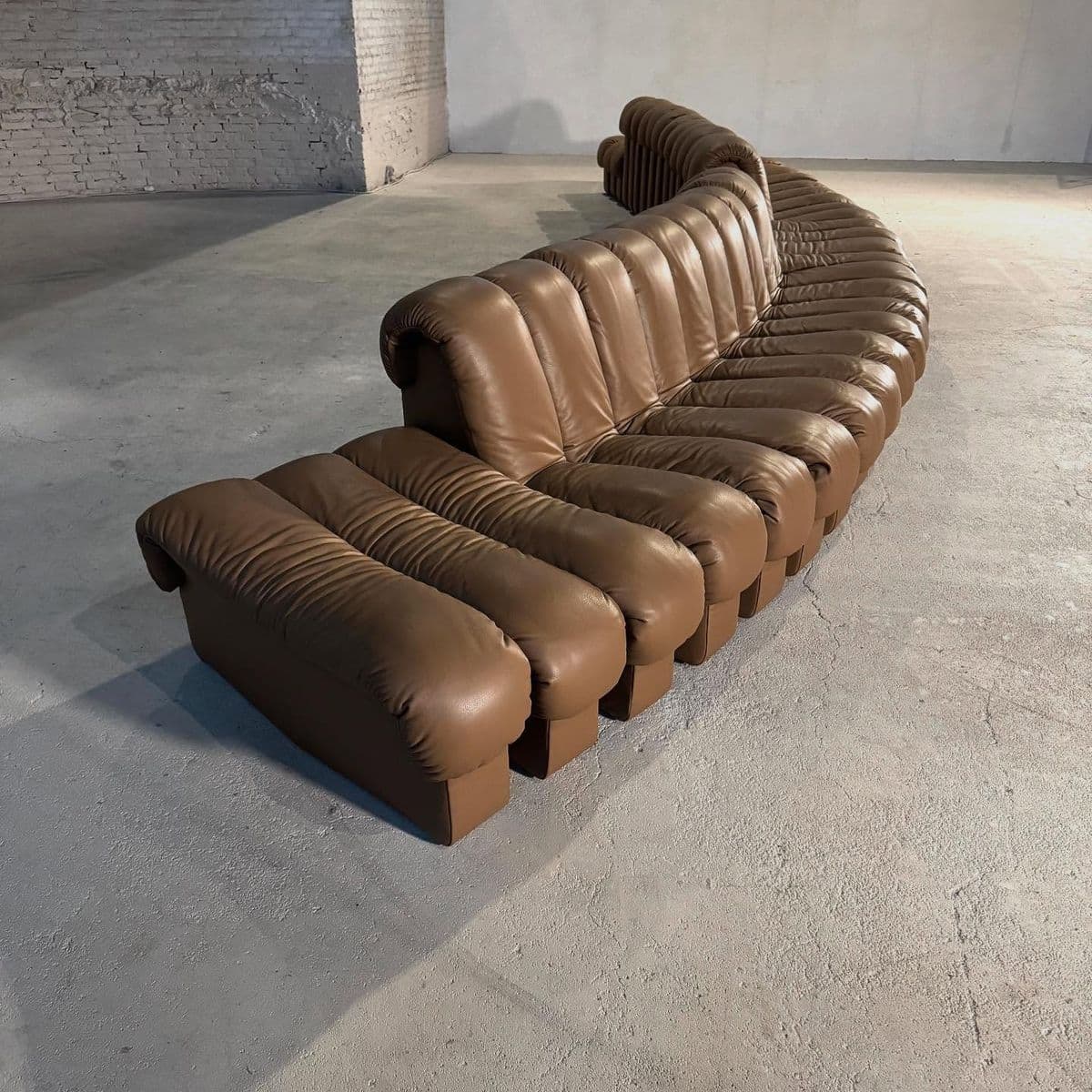 A modular, curved sectional sofa upholstered in brown leather. The sofa features a distinctive design with vertical channel tufting on the seat and back cushions, creating a segmented appearance. It is composed of multiple individual units that form a large, L-shaped seating arrangement with an additional projecting section. — 3D model