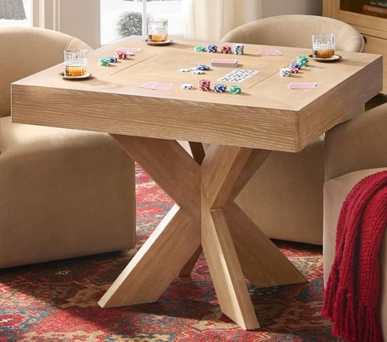 A modern square game table constructed from light oak wood, featuring a distinctive cross-pedestal base and integrated recesses on the tabletop for holding cards, poker chips, and beverages. The table is set up for a card game. — 3D model