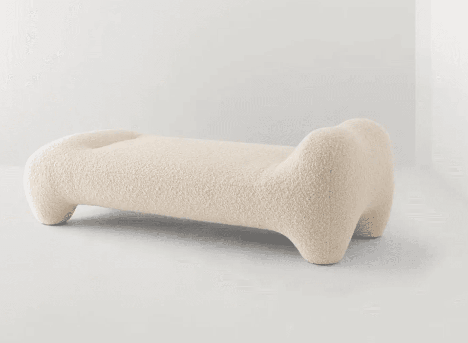 A long, low-profile, sculptural bench upholstered in a light, textured fabric, likely bouclé. It features organic, rounded forms with no hard edges, resembling an abstract bone or pebble shape. The design is minimalist yet impactful, intended as a statement piece. — 3D model