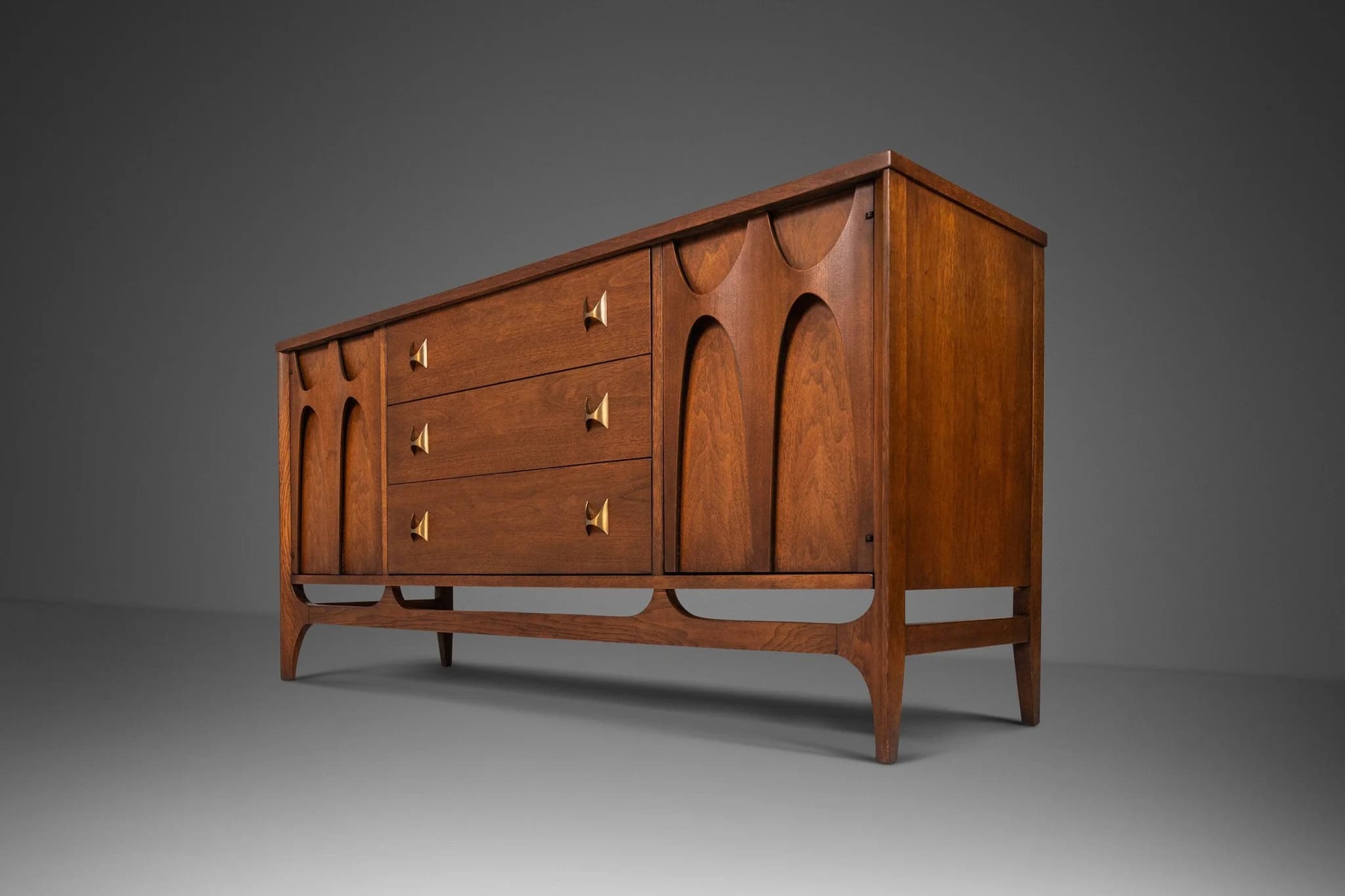 A long, wooden sideboard featuring three drawers with distinctive brass pull handles and two cabinet doors with carved, arch-like designs. It stands on an integrated, sculpted wooden base, typical of mid-century modern design. — 3D model