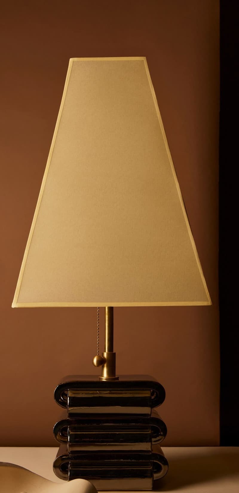 A modern table lamp featuring a stacked, polished chrome base composed of three distinct, curved, rectangular sections. The base supports a brass-finished neck and finial, from which a pull chain extends for operation. Crowning the lamp is a beige, tapered rectangular fabric shade, which complements the metallic base. The overall design suggests a contemporary aesthetic. — 3D model