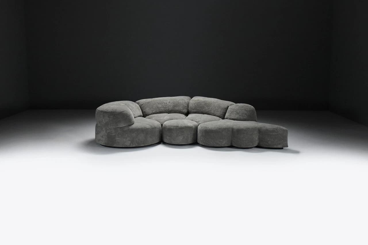 A large, multi-part sectional sofa featuring soft, organic, rounded forms, upholstered in a textured grey fabric. The sofa comprises several modules including corner pieces, armless central sections, and an end module that functions as an extended footrest or chaise. The overall design emphasizes comfort and a flowing, contemporary aesthetic. — 3D model