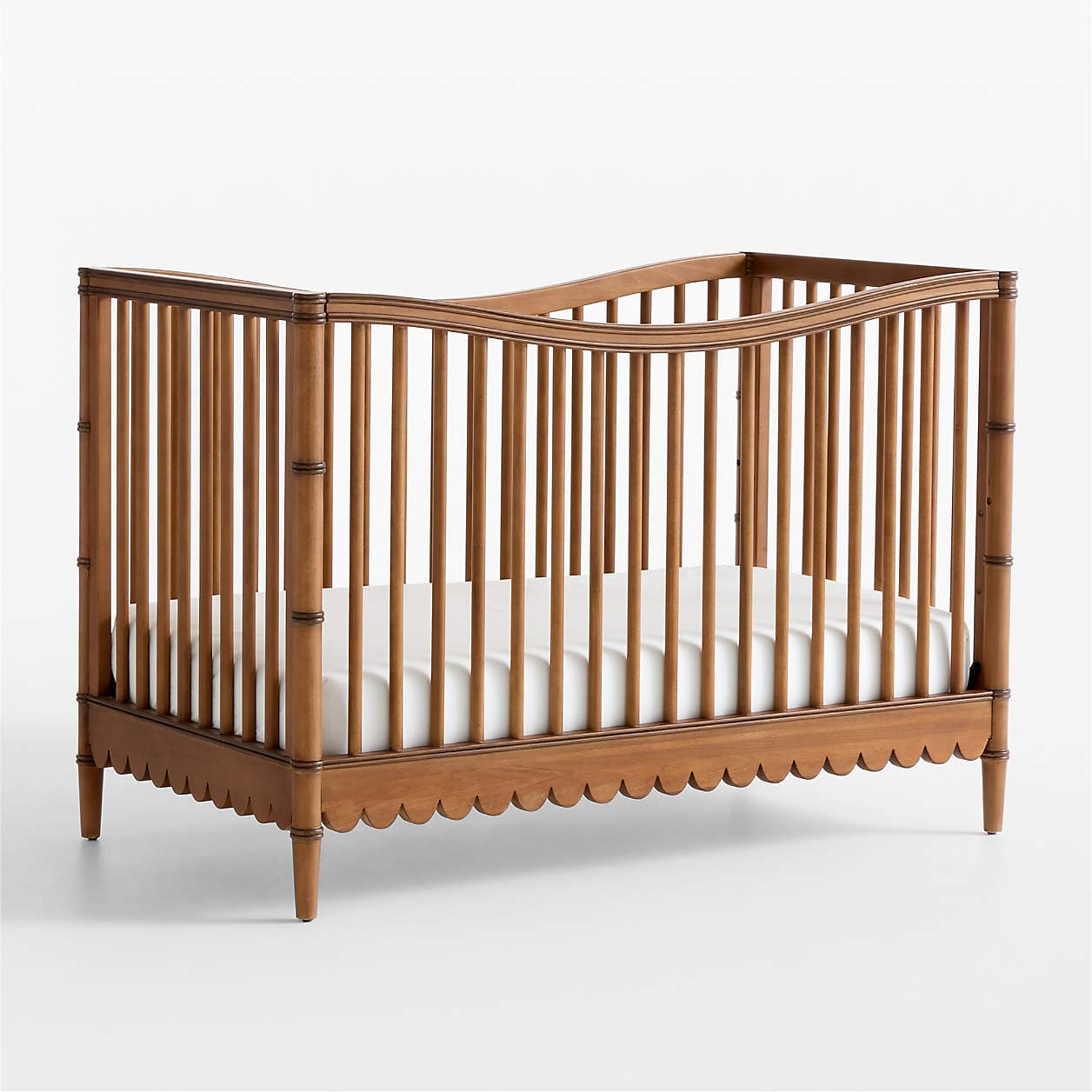A wooden crib with a scalloped bottom edge and turned legs. The crib features vertical spindles and a slightly curved top rail. A white mattress is present inside. — 3D model