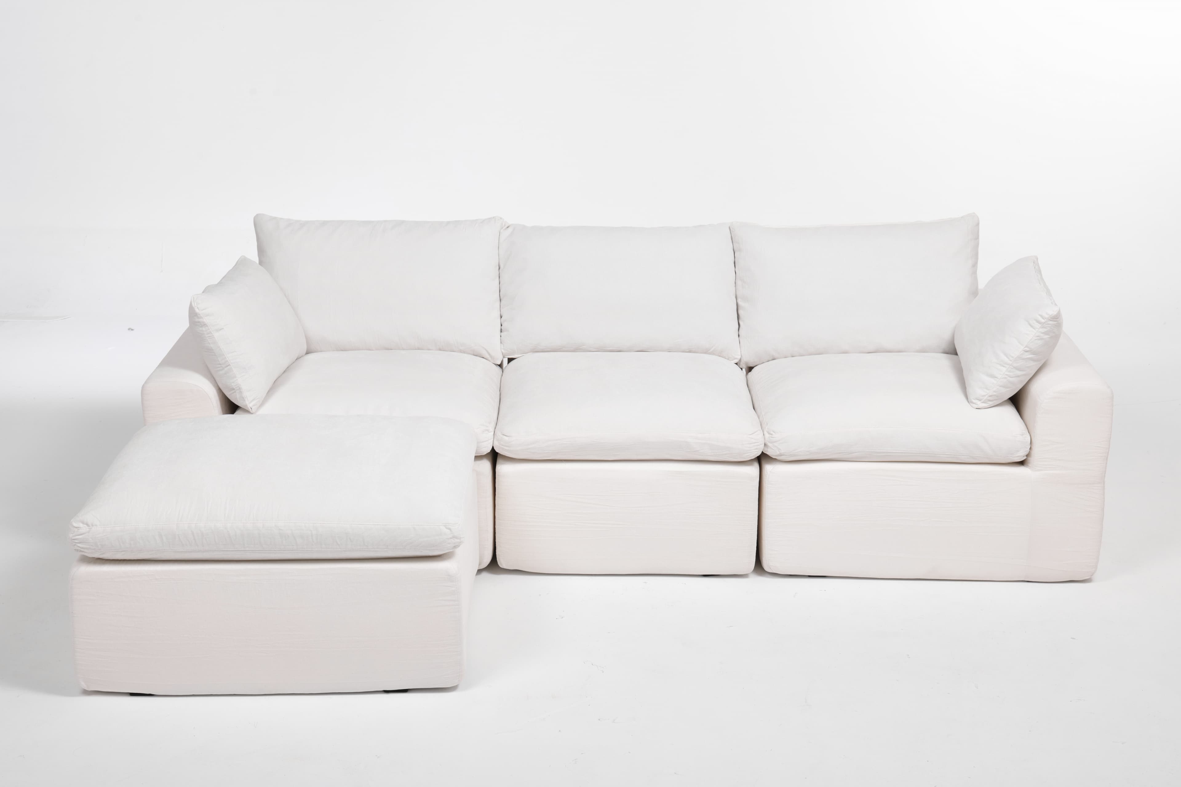 A modular sectional sofa with an attached ottoman. The sofa features deep, plush cushions and a low profile. It is upholstered in a textured white fabric. The configuration shown includes a corner section, two seat cushions, and an ottoman. The design is contemporary and emphasizes comfort. — 3D model