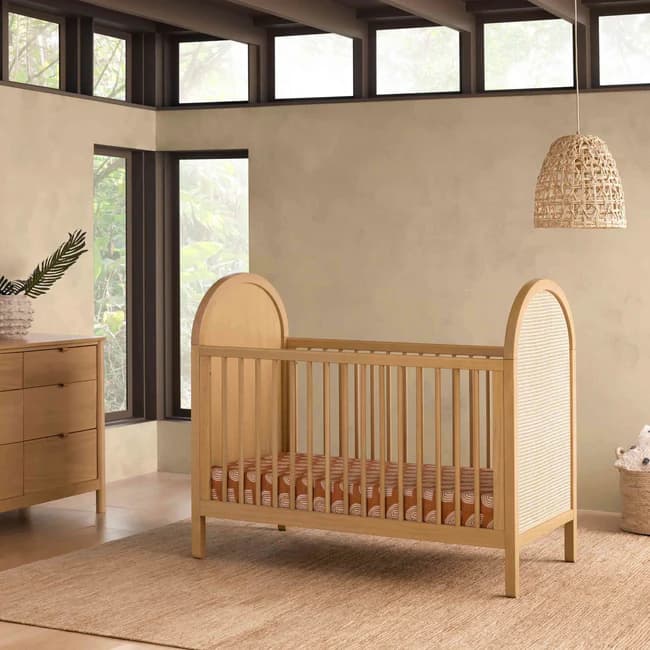 A wooden crib with a curved headboard and footboard. The sides feature vertical wooden slats, and one end panel is a woven cane material. A patterned mattress is visible inside. — 3D model