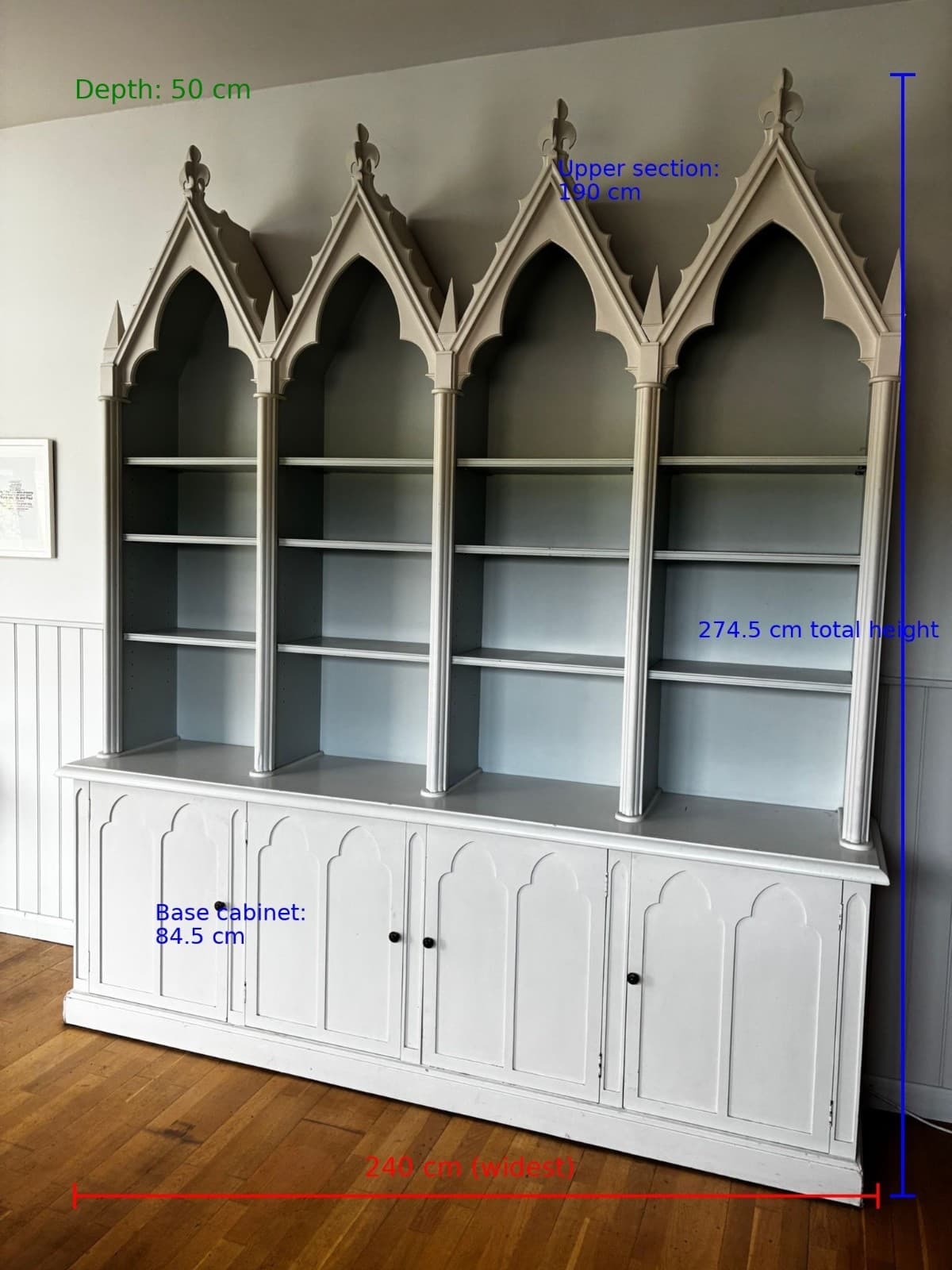 A tall, multi-sectioned bookcase hutch featuring a distinct Gothic Revival architectural style. The upper section consists of four open shelving bays, each topped with a prominent pointed arch and detailed tracery, culminating in decorative finials. The lower section provides enclosed storage with four paneled cabinet doors, each equipped with a small, dark spherical pull. The entire unit is rendered in a uniform light gray finish, suggesting painted wood or a similar material. The hutch sits on a base with a simple plinth. — 3D model