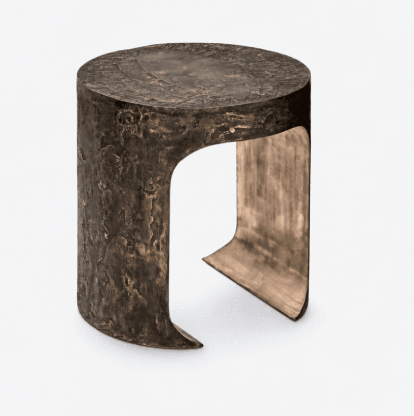 A cylindrical side table or stool crafted from patinated bronze or a similar metal, featuring a heavily textured, organic surface on its exterior. The design incorporates a single, wide, curved cut-out that forms a sculptural support, with the interior surface exhibiting a smoother, subtly brushed metallic finish. The top surface of the table also presents a nuanced, low-relief pattern. — 3D model