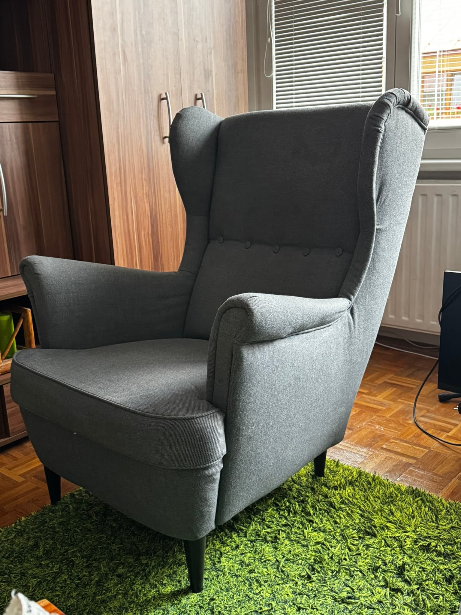 A contemporary wingback armchair upholstered in a textured grey fabric. The chair features a tall, slightly curved back with pronounced 'wings' at the top, a series of five decorative buttons forming a line down the center of the backrest, and sloped, rolled armrests. It has a removable seat cushion and stands on four slender, tapered dark-stained wooden legs. The overall design combines classic wingback elements with a modern, streamlined aesthetic. — 3D model