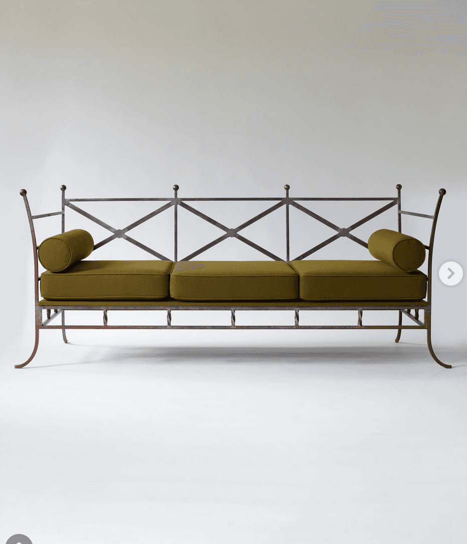 A three-seater sofa with a dark metal frame and green upholstered cushions. The backrest features a decorative criss-cross pattern with finials on top. Two cylindrical bolster cushions are placed at either end of the sofa. The frame has an industrial or antique aesthetic, with visible joins and a slightly weathered appearance. — 3D model