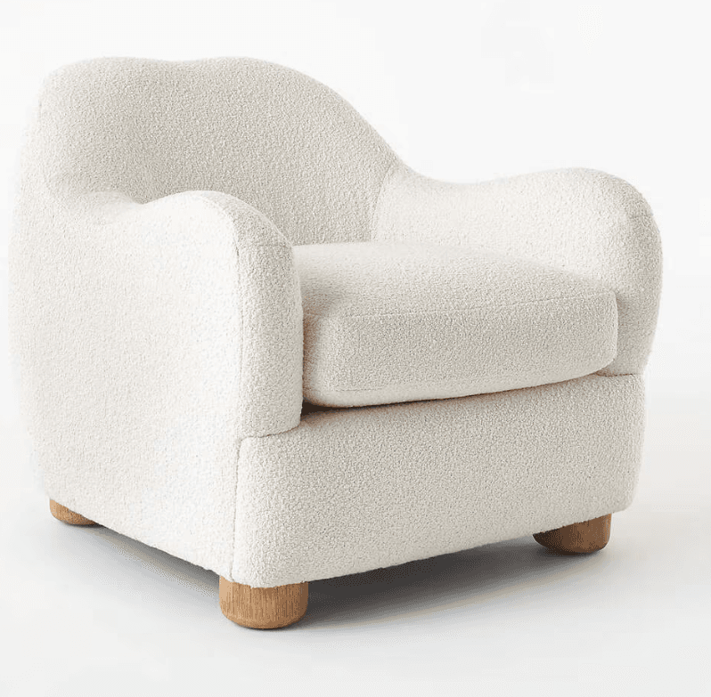 A contemporary armchair fully upholstered in a cream-colored bouclé fabric, featuring a soft, rounded silhouette. The chair has a deep seat with a removable cushion and gently curved armrests and backrest, all contributing to a plush and inviting appearance. It stands on four short, cylindrical, light-toned wooden legs. — 3D model