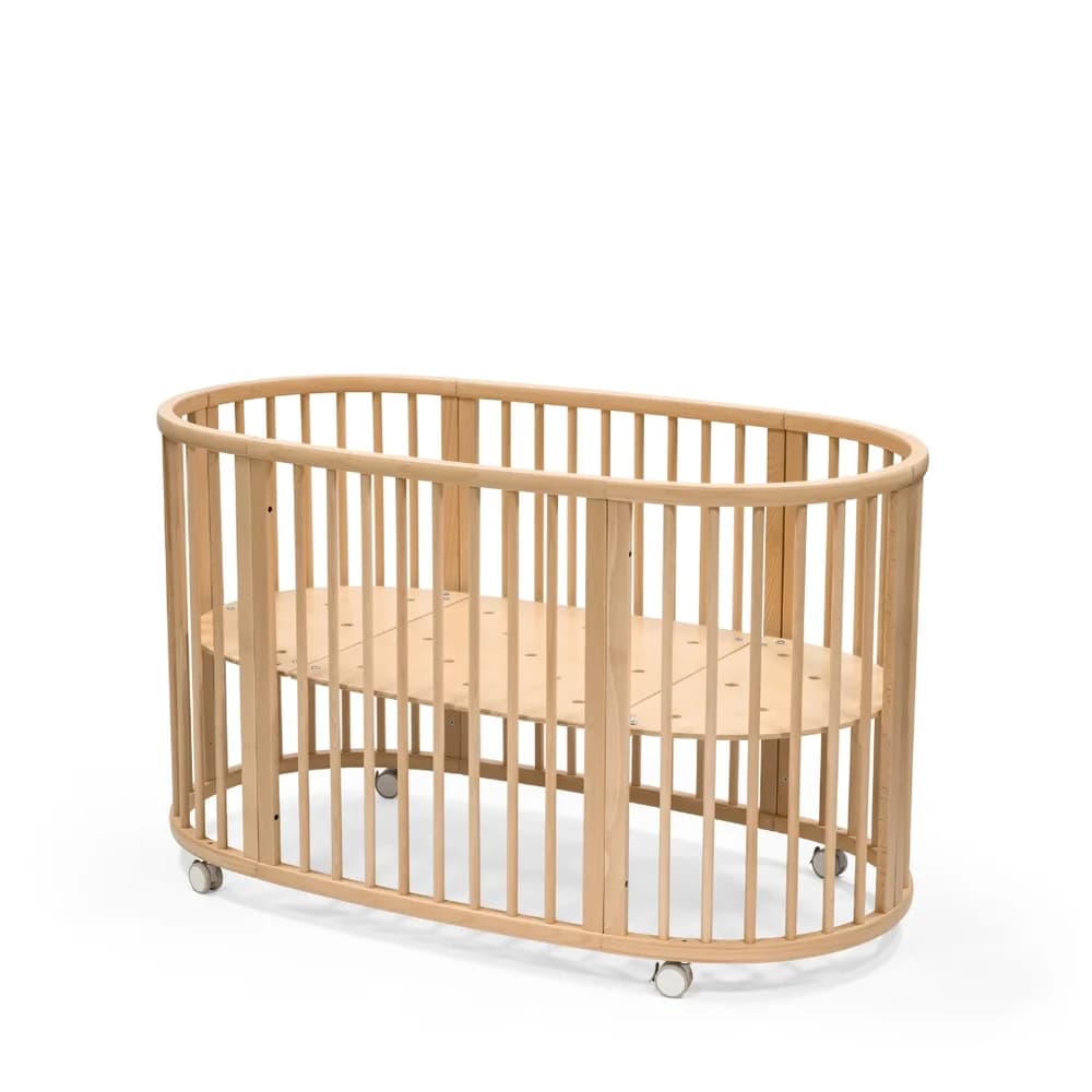 A natural wood, oval-shaped baby crib featuring vertical slats for safety and visibility, and small casters on the base for mobility. The mattress support is a slatted wooden panel. — 3D model