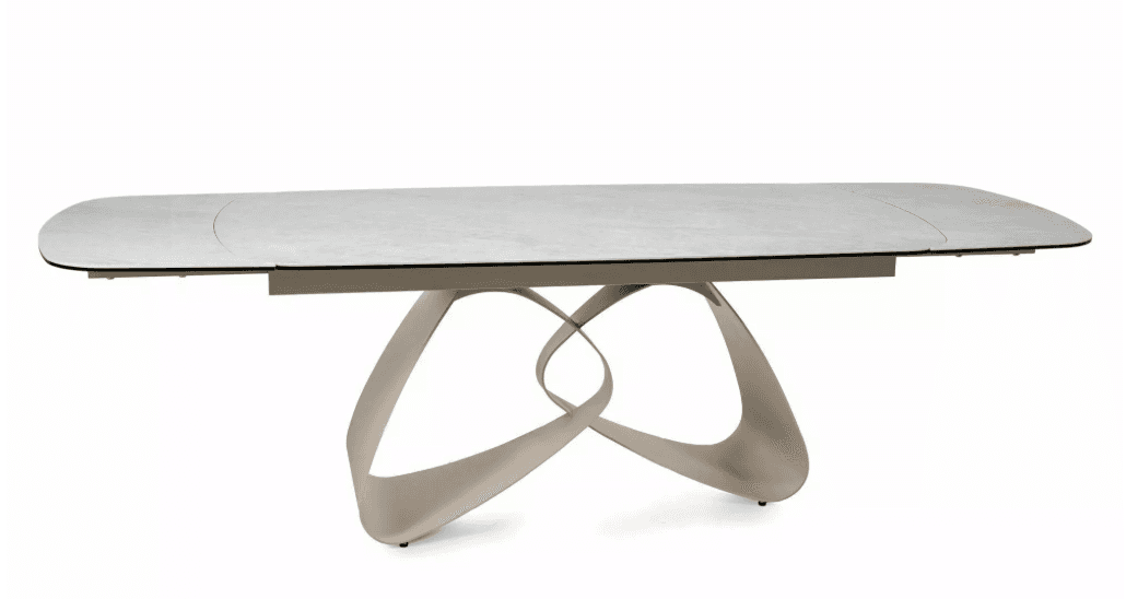 A modern dining table with a rectangular, elongated tabletop featuring softly rounded corners. The tabletop has a subtle texture and appears to be a light, neutral color, possibly white or light gray. It is supported by a sculptural base composed of interlocking, fluid shapes that create an abstract, organic form. The base has a metallic or polished finish, reflecting light and adding a dynamic visual element. The overall design suggests contemporary aesthetics. — 3D model
