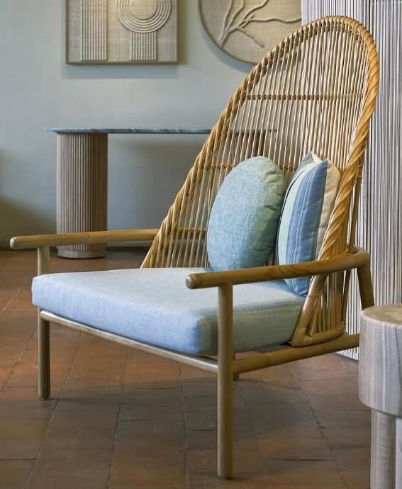A contemporary lounge chair featuring a light-toned solid wood frame and an intricately woven natural rattan backrest. The chair is equipped with a thick, light blue fabric seat cushion and two decorative throw pillows, one round and solid light blue, and one rectangular with blue and white stripes. The design emphasizes natural materials and a relaxed aesthetic. — 3D model