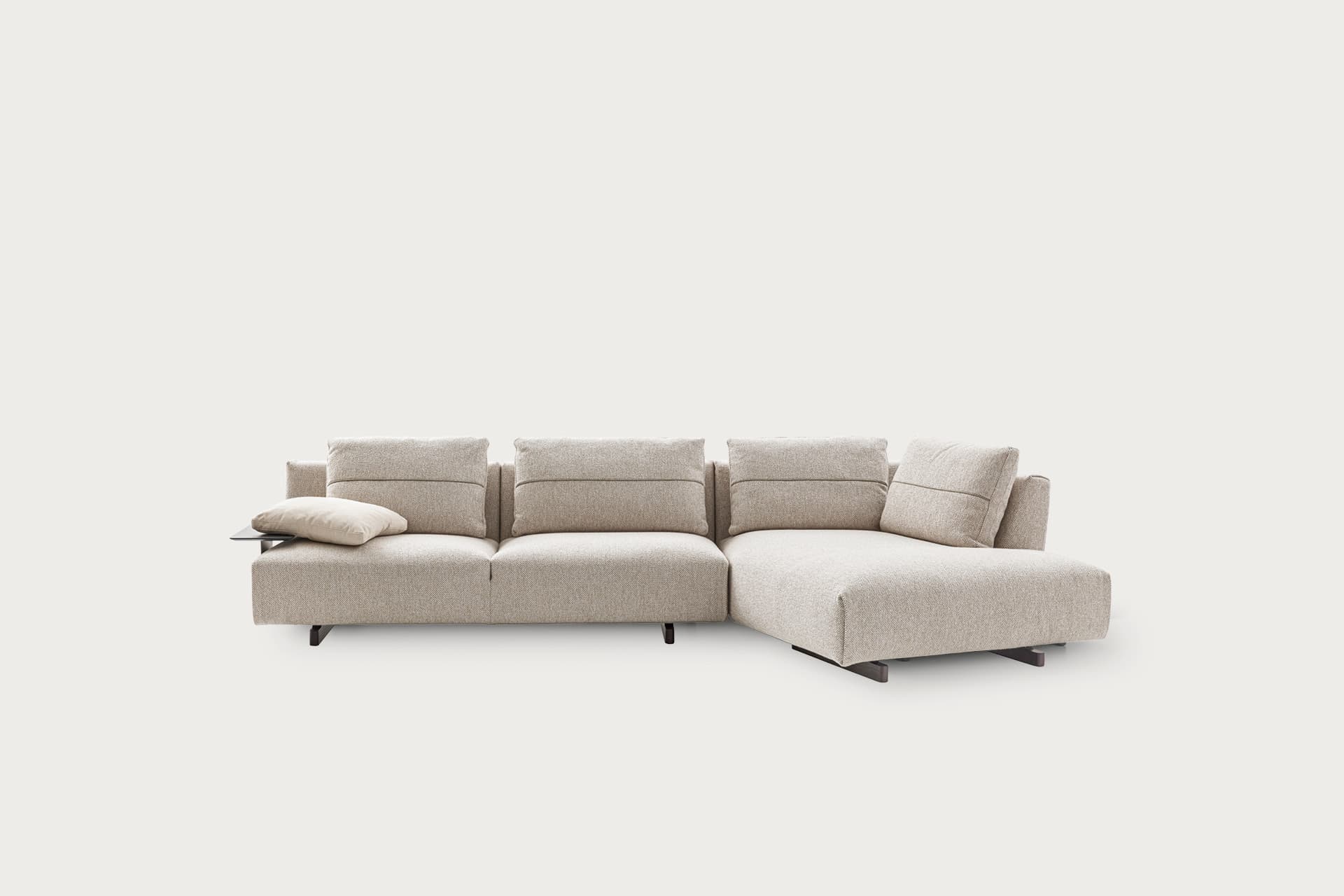 A contemporary modular sectional sofa featuring a chaise lounge component and an integrated side table on one end. The sofa is upholstered in a light, textured fabric, possibly a beige or oatmeal color, with a low-profile design. It consists of multiple segments with individual back cushions that feature horizontal stitching details, giving them a structured yet inviting appearance. The sofa is supported by slender, dark-colored, minimalist metal legs or a plinth base, contributing to its modern aesthetic. A light-colored throw pillow rests on the end module with the side table. — 3D model