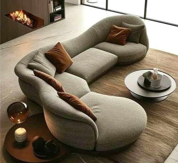 A modern, modular sectional sofa with a curved, organic design. The sofa features a light beige textured fabric upholstery and a dark brown wooden base. It includes several throw pillows in shades of brown and taupe. The configuration suggests it can be adapted to various room layouts. — 3D model