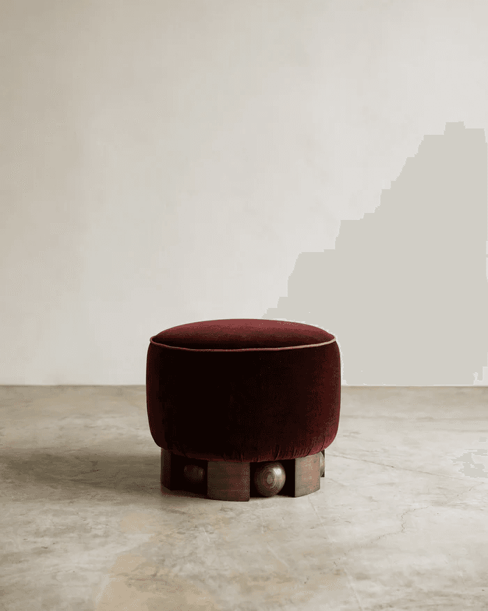 A round, upholstered ottoman with a deep red velvet fabric. The ottoman is characterized by a decorative piping along its top edge and a unique base constructed from dark wood elements, including carved spherical shapes and angular blocks arranged in a tiered pattern. The base provides a contrast to the soft upholstery of the seat. — 3D model