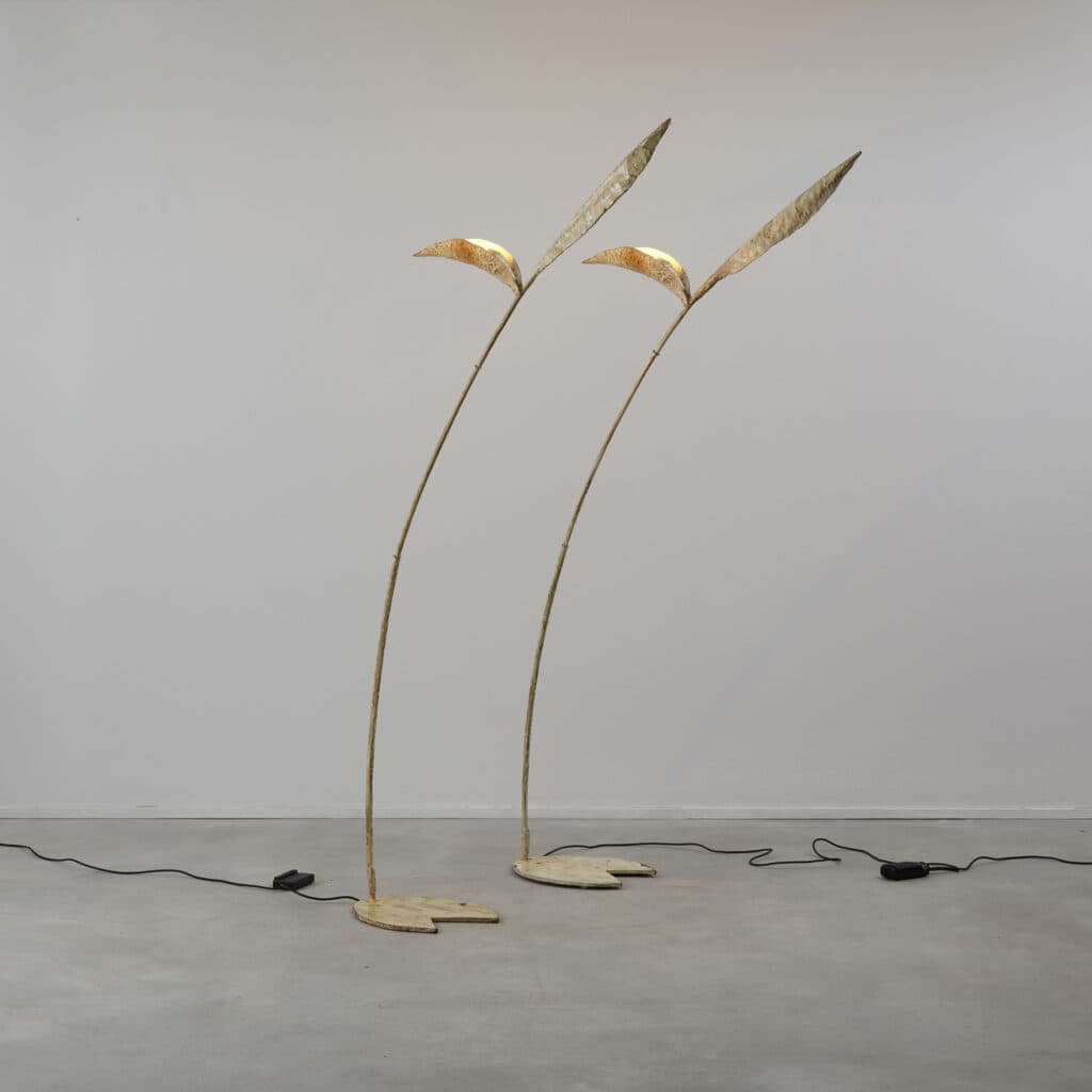 A tall, slender floor lamp with an organic, botanical design, featuring a distressed gold or bronze finish. The lamp has a round, flat base from which a curved stem extends upwards, culminating in two leaf-shaped elements that house the light sources. A black power cord with an inline foot switch extends from the base. — 3D model