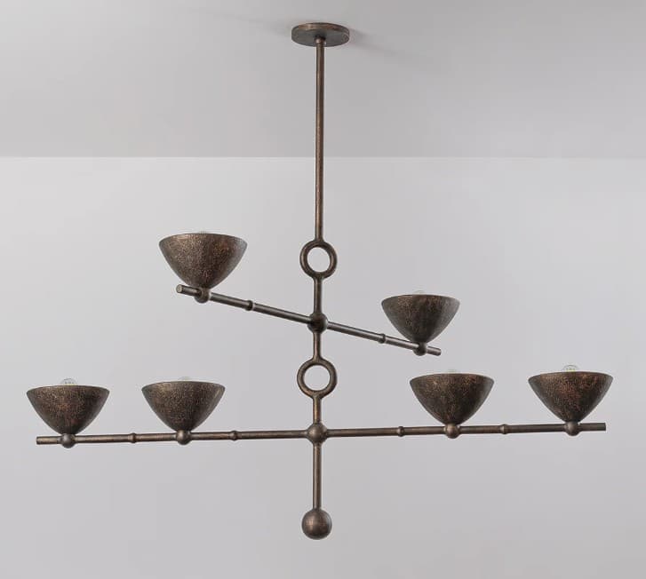 A six-light chandelier featuring a linear, branching design with a distressed bronze finish. The fixture hangs from a ceiling canopy via a single downrod. Its central vertical stem branches into two horizontal arms, with the upper arm supporting two light sources and the lower, longer arm supporting four. Each light source is housed within a small, bowl-shaped shade, pointing upwards. Circular accents are integrated into the central stem where the arms attach. The metal surface has a textured, aged appearance. — 3D model