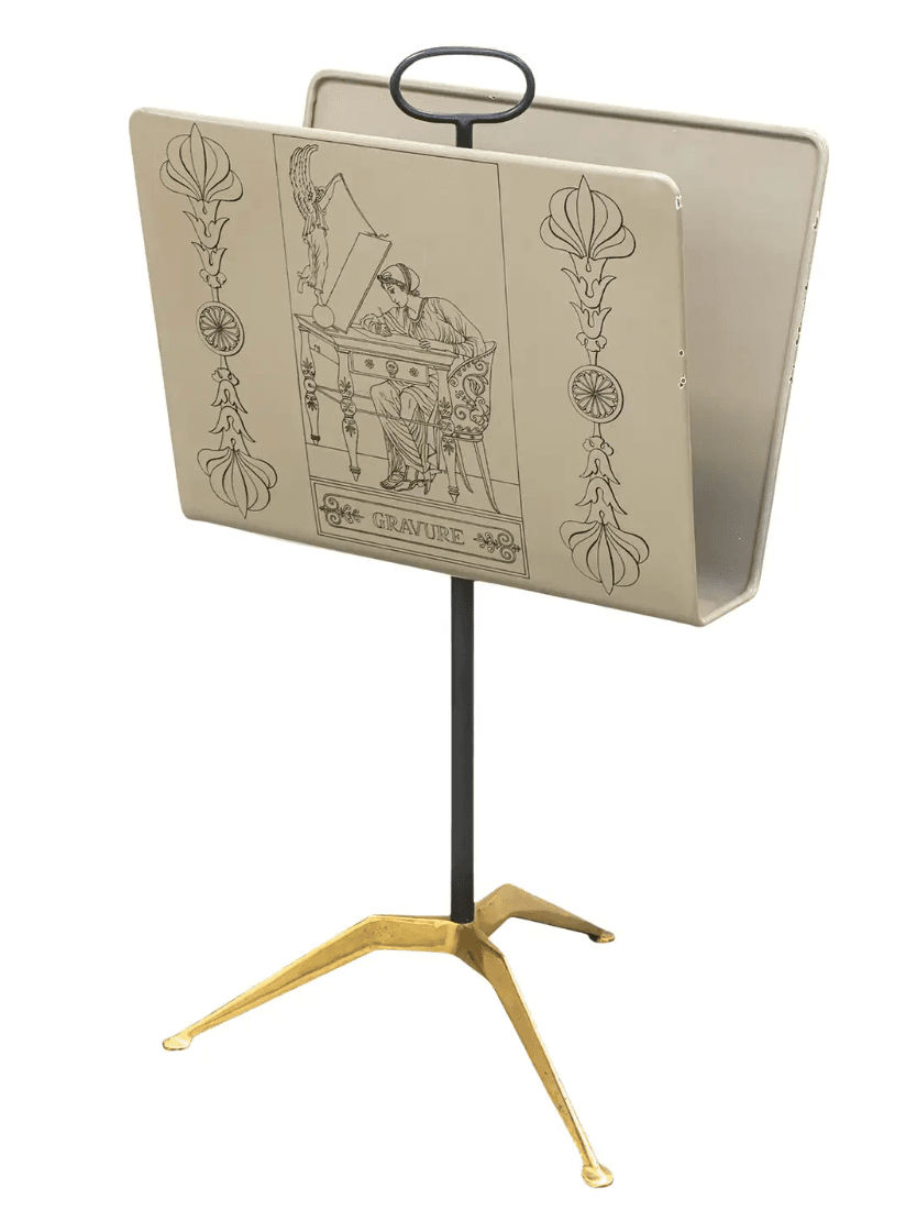 A freestanding magazine rack featuring a light-colored enameled metal trough adorned with black Art Deco-style line art illustrations. It is supported by a slender dark metal central pole and a distinctive three-legged brass base. A dark metal loop handle is integrated at the top for portability. — 3D model