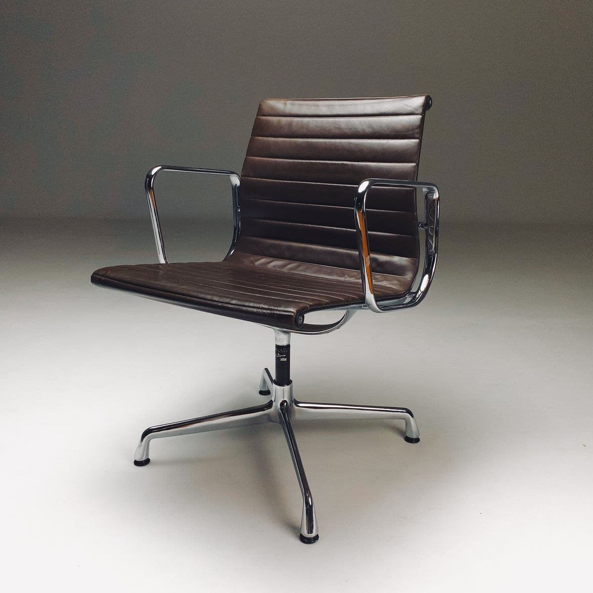 A mid-century modern desk chair with a polished aluminum five-star base and arms. The seat and back are upholstered in dark brown leather with horizontal channel stitching. The chair features a central pedestal that supports the seat and back assembly, and it appears to be on casters or glides for mobility. The design is minimalist and functional, characteristic of iconic mid-century design. — 3D model