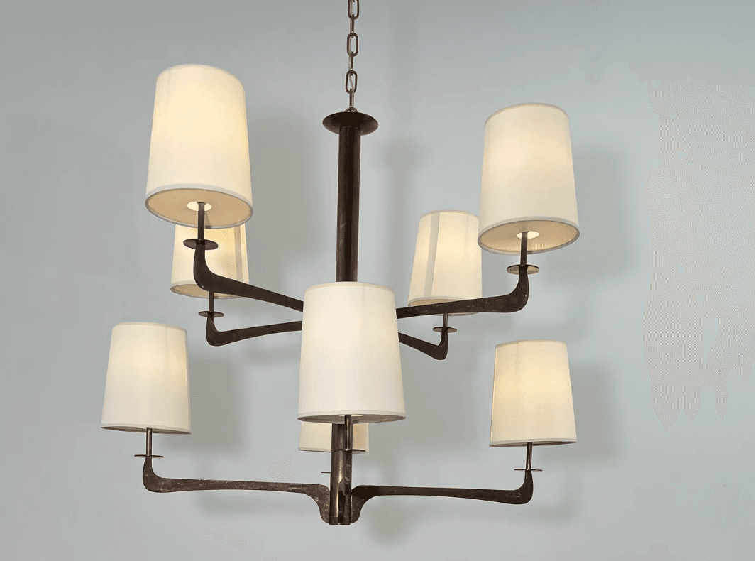 A modern multi-arm chandelier featuring a dark bronze-finished metal frame and eight tapered, drum-style fabric shades. The central column extends from a chain suspension, with eight arms branching out, each supporting an upward-facing light socket covered by a cream-colored textile shade. The metalwork exhibits a rustic or aged patina, suggesting a blend of modern design with traditional material treatment. — 3D model