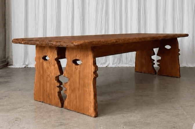 This is a substantial dining table crafted from solid wood, featuring a live-edge tabletop that retains the natural contour of the tree. The most distinctive element is its base, which consists of two thick, rectangular wood slabs that are intricately carved with abstract human face profiles. When placed together, the two halves of the base interlock, creating a cohesive and sculptural support system for the table. The wood exhibits a warm, medium brown tone with visible grain patterns and a rustic, unfinished or lightly finished texture, highlighting its organic character. — 3D model