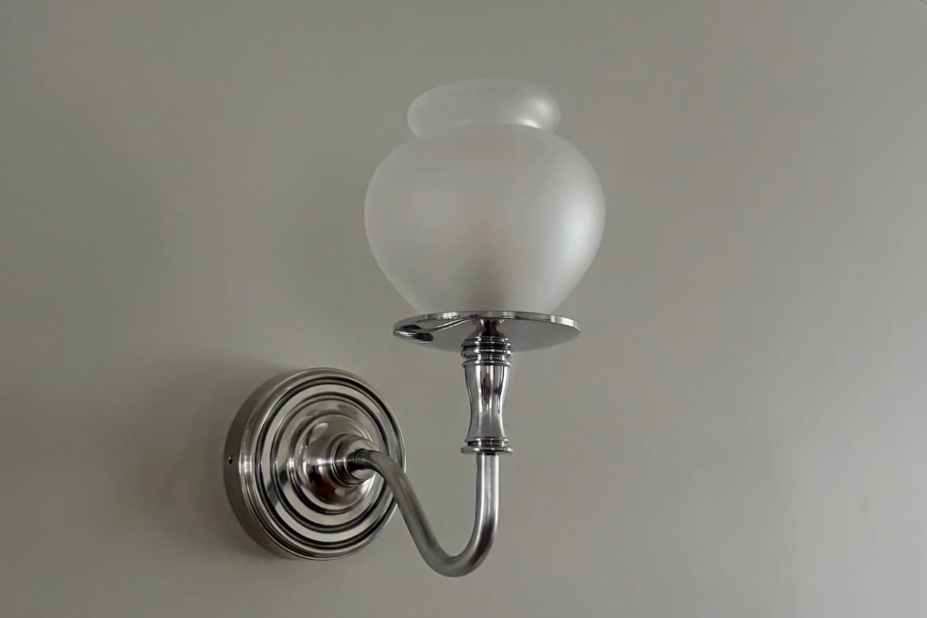 A polished nickel or chrome wall sconce featuring a frosted glass shade in an urn-like shape. The arm is gently curved, extending from a circular, fluted backplate, providing ambient or accent lighting. — 3D model
