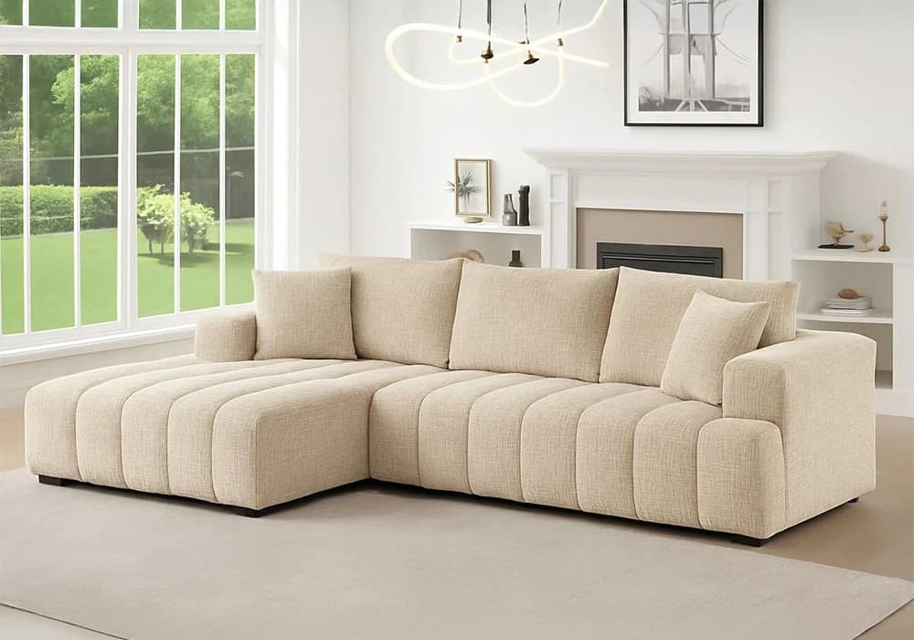 A modern L-shaped sectional sofa with a fixed left-hand chaise, upholstered in a light-colored woven fabric. The base and chaise feature distinctive vertical channel tufting. It includes multiple loose back cushions and accent pillows, supported by discreet black block feet. — 3D model
