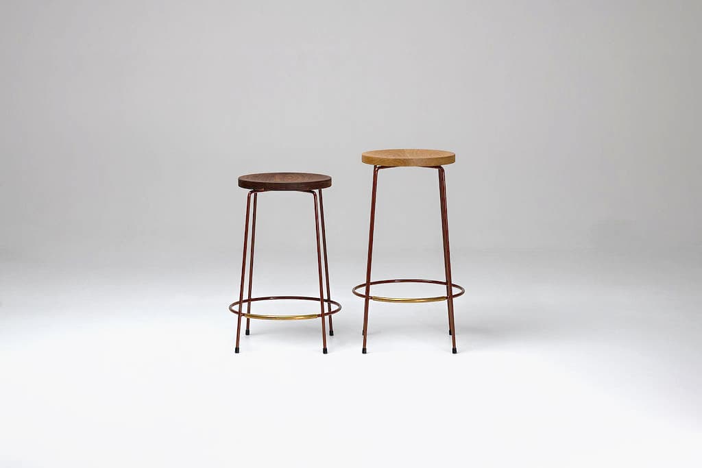 A minimalist bar stool featuring a round, natural oak wood seat and a slender four-legged metal base. The legs are a dark reddish-brown hue, tapering slightly towards the floor, and are reinforced with a circular brass-toned metal footrest. — 3D model