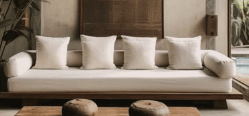A modern, low-profile sofa featuring a dark wooden platform base and light cream-colored upholstery. It includes a single long seat cushion, multiple square back cushions, and cylindrical bolster pillows serving as armrests. — 3D model