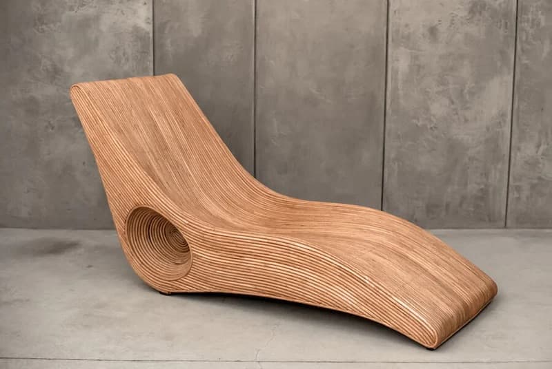 A contemporary chaise lounge with a sinuous, flowing design. The piece is constructed from layered, bent wood, creating a ribbed texture along its surface. It features an integrated circular opening near the base of the backrest, adding an unconventional aesthetic detail. The overall form is ergonomic, designed for reclining. — 3D model