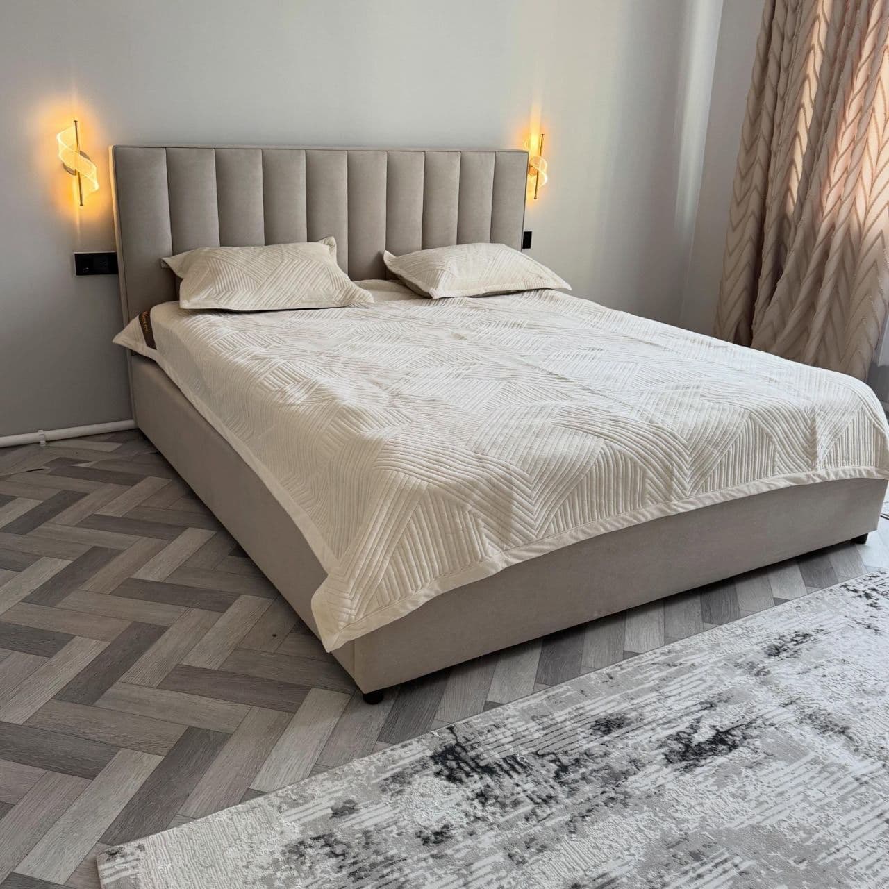 A modern upholstered platform bed featuring a tall headboard with vertical channel tufting. The entire bed frame, including the headboard and side rails, is covered in a neutral-toned fabric, likely beige or light gray. The bed is presented with a white mattress, two matching pillows, and a textured white duvet/quilt. It has a low-profile design, sitting on small, recessed feet. — 3D model