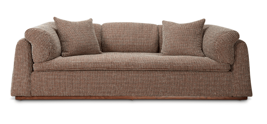A three-seater sofa with a low profile and rounded arms. It features a patterned upholstery in shades of brown and white, reminiscent of a houndstooth or small check. Two square throw pillows with matching upholstery are placed on the sofa. The sofa rests on a visible wooden base. — 3D model
