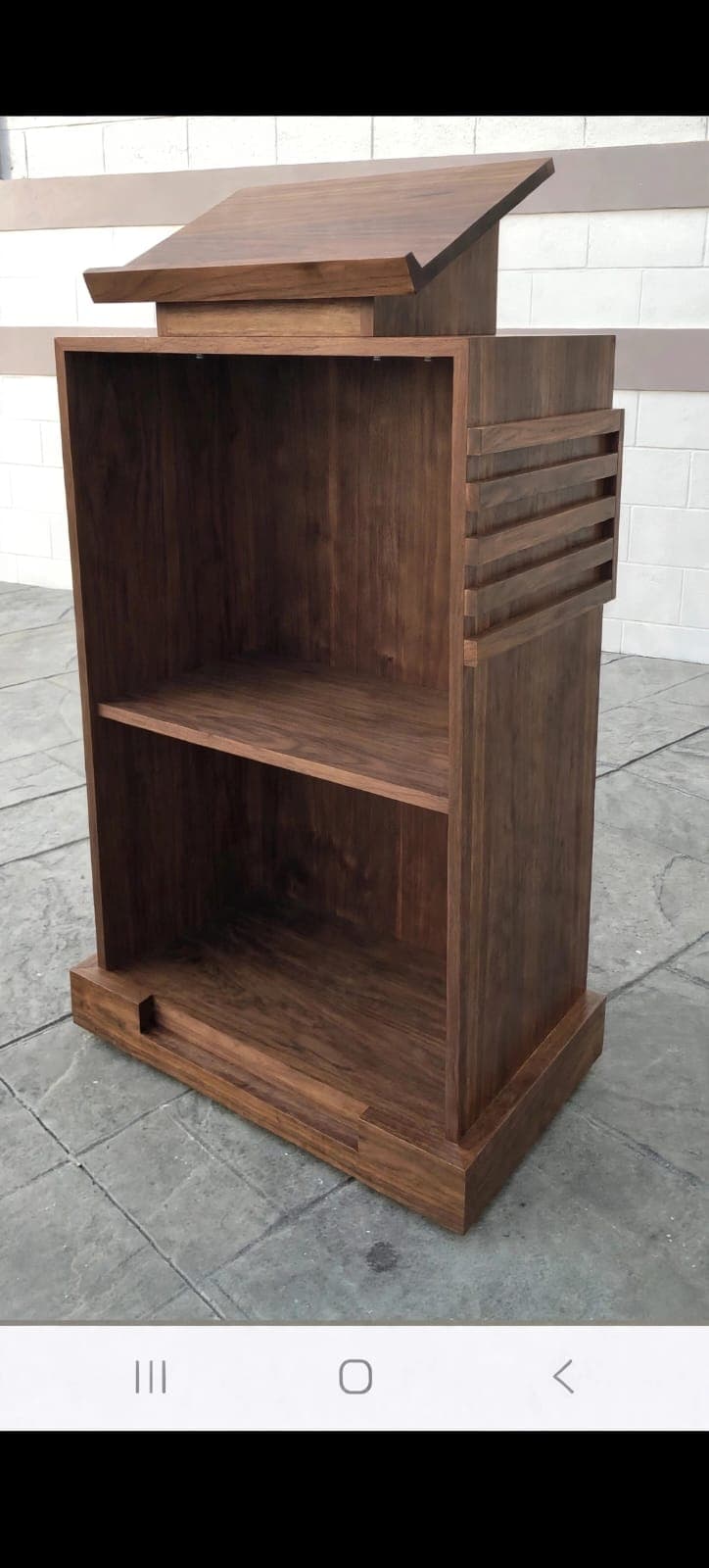 A modern, freestanding wooden lectern featuring a dark stained finish. It has a slanted reading surface with a small lip, supported by a block base. The body of the lectern contains open shelving compartments for storage. One side panel is adorned with horizontal slatted detailing, while the other side is plain. The base of the unit is slightly recessed, creating a plinth-like appearance. — 3D model