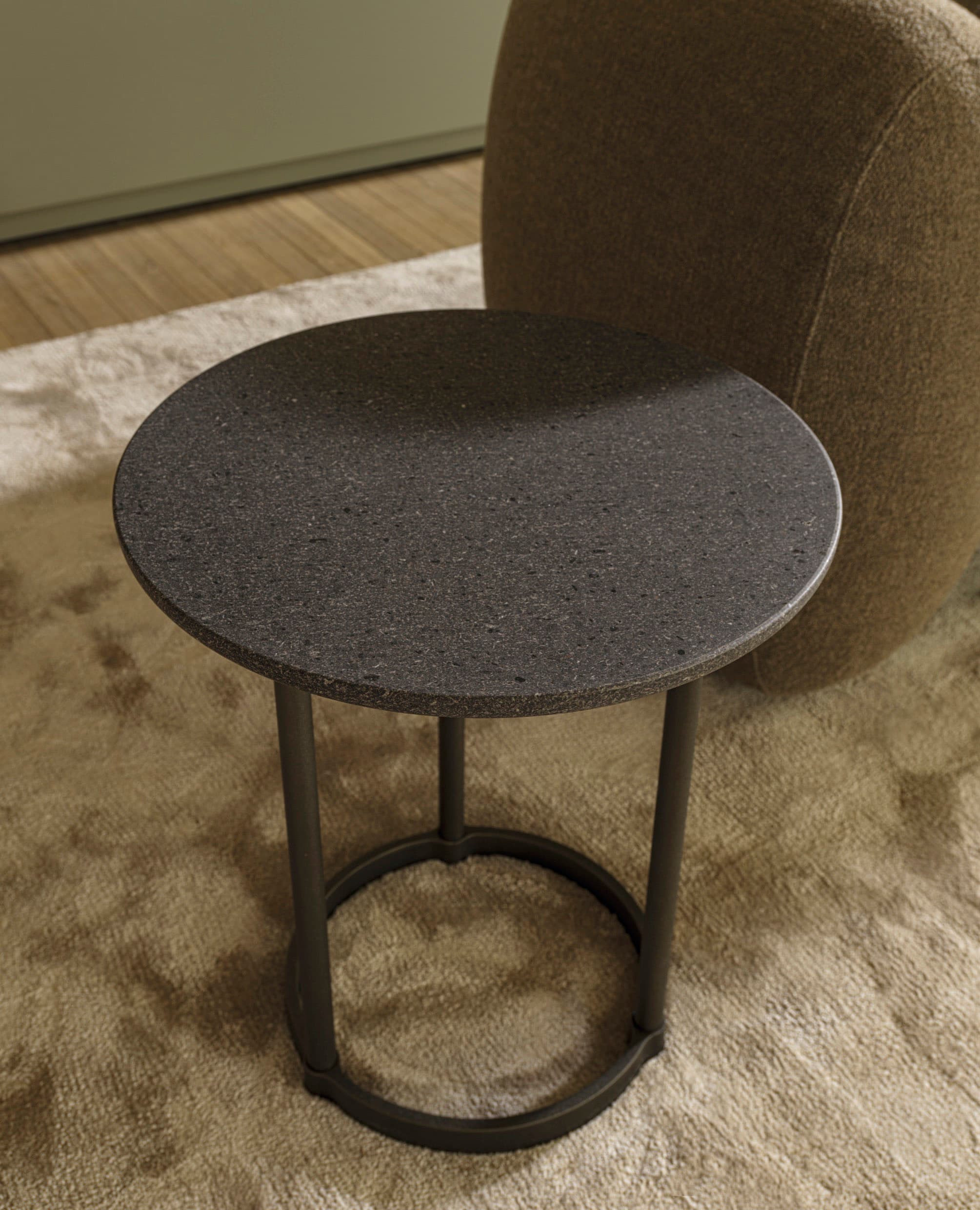 A small, round accent table featuring a dark, granular-textured top and a minimalist metal base with three slender legs connected by a circular stretcher. — 3D model