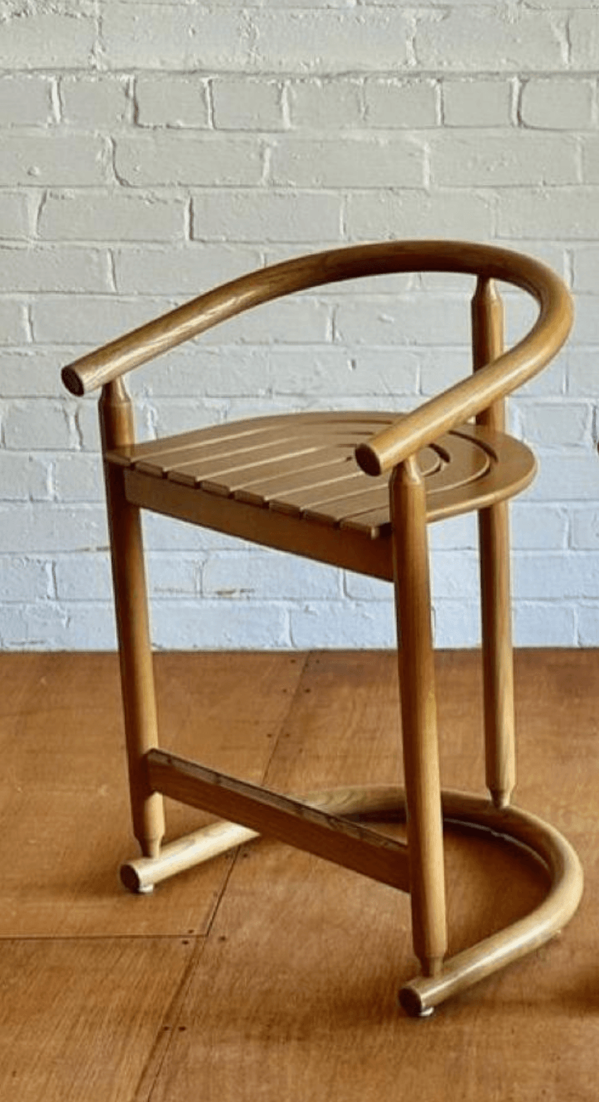 A contemporary bar stool made from light-toned wood, featuring a slatted seat and a continuous semi-circular backrest that doubles as armrests. It has four tapered legs and a curved footrest. — 3D model