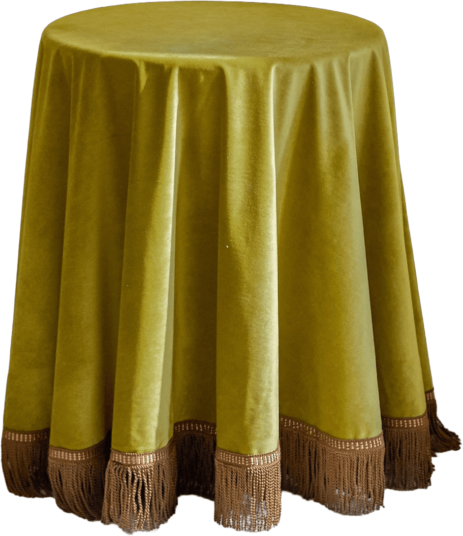 A round table completely covered by a floor-length, olive green velvet tablecloth. The tablecloth features a decorative fringe trim in a contrasting brown color along its bottom edge. The velvet fabric displays a soft sheen and drapes in gentle folds around the table, obscuring the base. The fringe is segmented by a patterned band just above the tassels. — 3D model