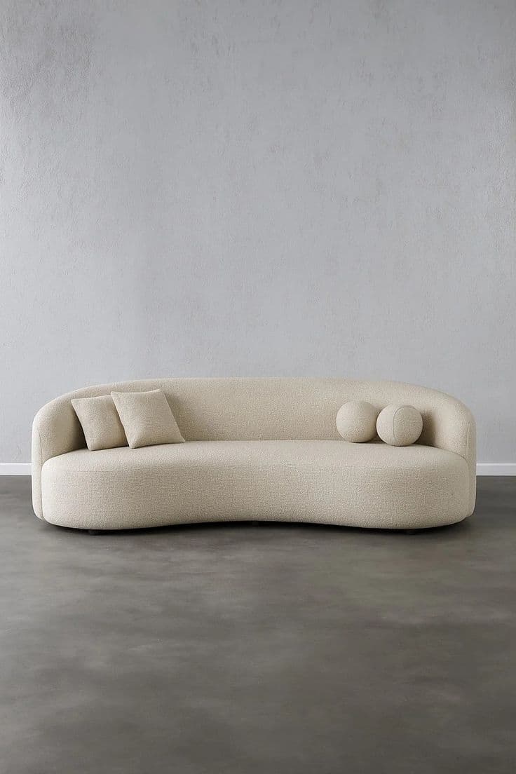 An asymmetrical curved sofa upholstered in a light beige, subtly textured fabric. It features a continuous, low-profile backrest and armrest, flowing into a single, organic form. The sofa is accompanied by two small rectangular throw pillows and two spherical decorative cushions, all in matching upholstery. — 3D model